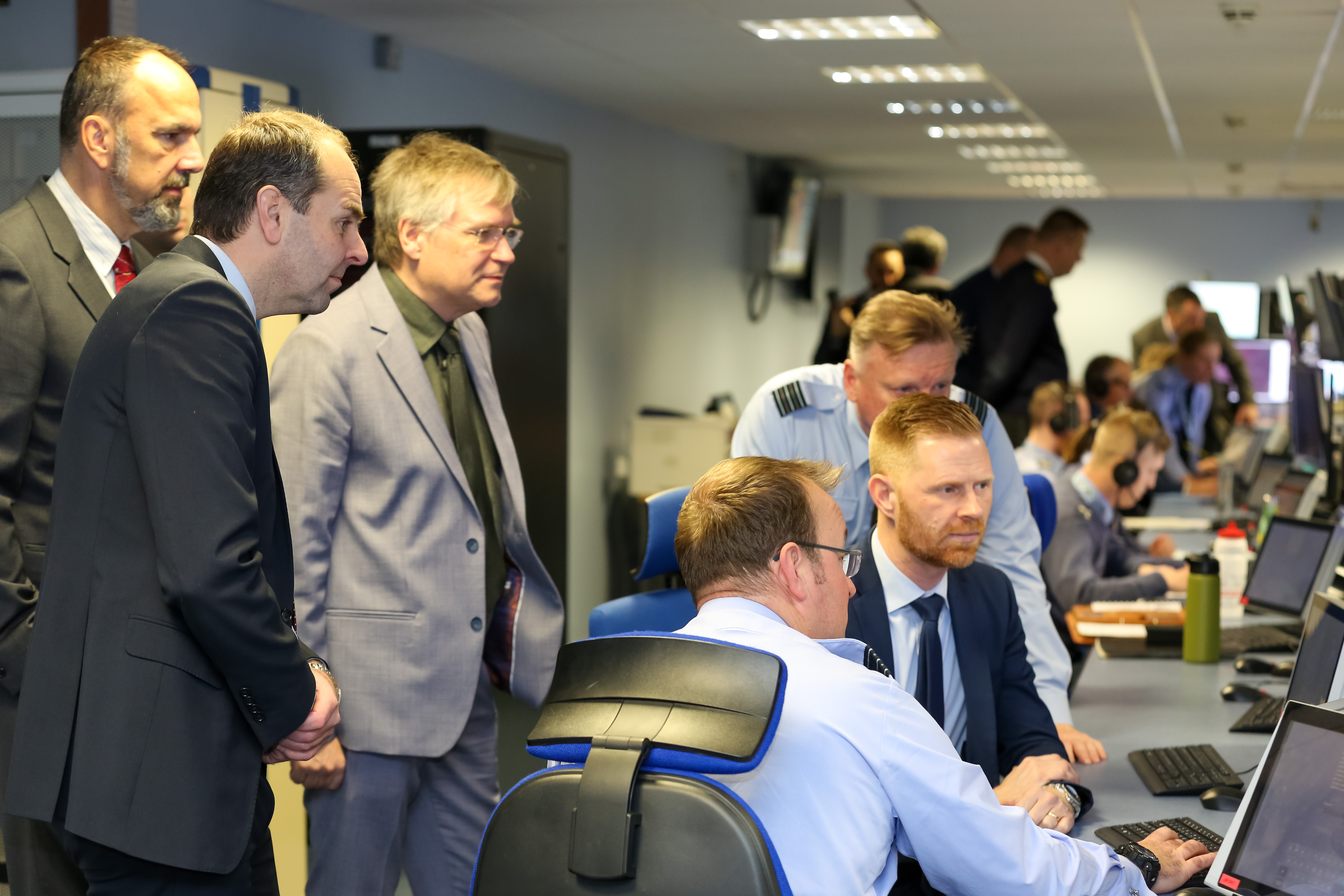 Image shows RAF aviators and civilians standing with discussion with others sitting at computer desk.